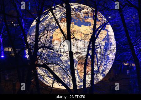 Brescia - Italia. 15. Februar 2023: Light is Life (Festa delle Luci A2A), „Floating Earth“ von Luke Jerram inmitten von blau beleuchteten, blattlosen Ästen von Bäumen Stockfoto
