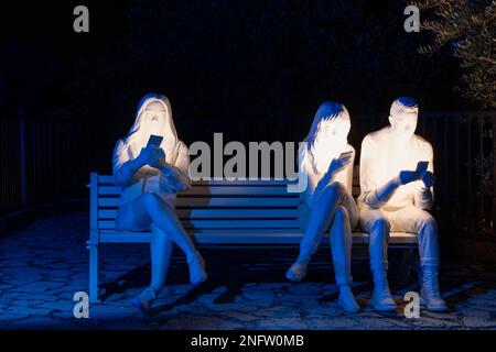 Brescia - Italia. 15. Februar 2023: Light is Life (Festa delle Luci A2A), beleuchtete menschliche Figuren, die Mobiltelefone halten und auf einer Bank sitzen Stockfoto