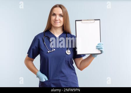 Eine Ärztin, die ein Klemmbrett mit leerem weißem Papier auf blauem Hintergrund hält Stockfoto