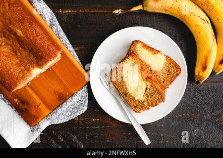 Scheiben mit Sahne, Käse gefülltes Bananenbrot auf einem weißen Teller: Scheiben von Bananenbrot mit reifen Bananen im Hintergrund Stockfoto