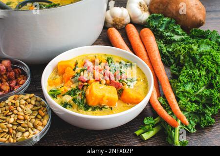 Süßkartoffelsuppe in einer Schüssel und holländischer Ofen mit Garnierungen: Cremige Gemüsesuppe garniert mit gebratenem Pancetta und gerösteten Pepitas Stockfoto