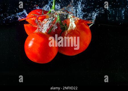 Frische rote Tomate, die ins Wasser fällt, mit Wasserspritzer und Luftblasen auf schwarzem Hintergrund. Stockfoto