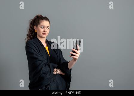 Lockig-haarige emotionale Teenager Frau in einem schwarzen Mantel im Gespräch per Videoanruf auf dem Smartphone, isoliert auf grau Stockfoto