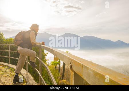Frau mit Rucksack lehnt sich auf ein Geländer und genießt den Panoramablick über Locarno und die Berge an einem nebligen Tag in Tessin, Schweiz. Stockfoto