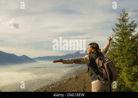 Frau mit Rucksack und ausgestreckten Armen Genießen Sie einen Panoramablick über Locarno mit Wolkenlandschaft in Ticino, Schweiz. Stockfoto