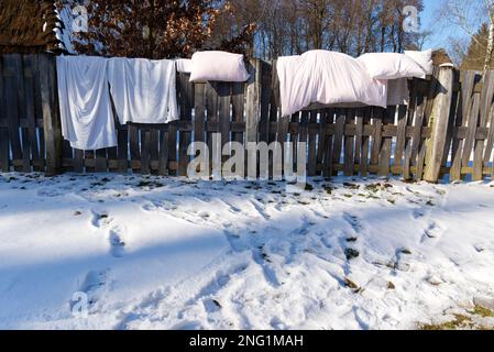 Weiße Bettwäsche wird im Winter auf einem alten Holzzaun in einem schneebedeckten Landgarten getragen Stockfoto