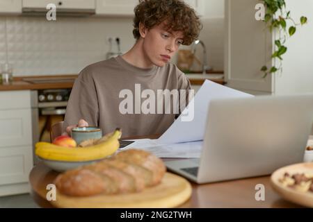 Porträt eines jungen, lockigen männlichen Studenten, der ein hausgemachtes Frühstück am Tisch serviert und sich auf die Prüfungsvorbereitungen konzentriert. Stockfoto