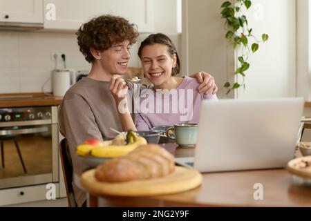 Nahaufnahme von jungen Inhalten fröhliches weißes Paar, das gesundes Essen zum Frühstück isst, lächelnd lächelt, sich einen Film auf dem Laptop ansieht. Stockfoto