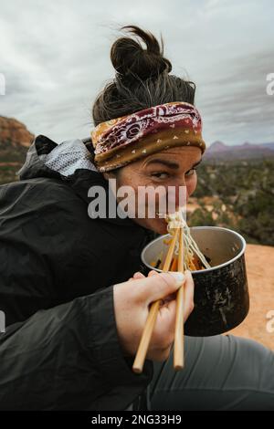 Junge, glückliche, wunderschöne weiße Frau, die 30s Lächeln vor der Kamera hat und Ramen aus dem Campingtopf mit Essstäbchen in Sedona, Arizona, USA, isst Stockfoto