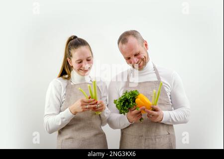 Glückliche attraktive junge Frau in Schürze, die frischen Salat zubereitet, mit fröhlichem bärtigen Ehemann in der Küche. Lächelndes, liebevolles Familienpaar, verheiratetes Paar, das zu Hause gesundes Essen kocht Stockfoto