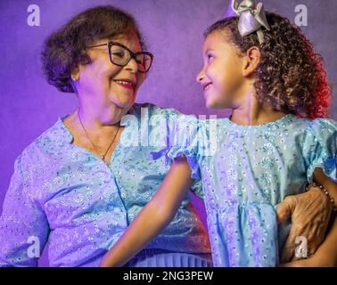 Studioporträt von Großmutter mit Enkelin in wunderschönen blauen Kleidern mit violetter Beleuchtung im Hintergrund Stockfoto