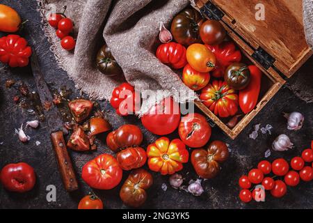 Viele verschiedene Arten, Formen und Größen von Tomaten in einer alten Holzkiste und auf einer dunklen Oberfläche, flach liegend, Draufsicht. Erntekonzept Stockfoto