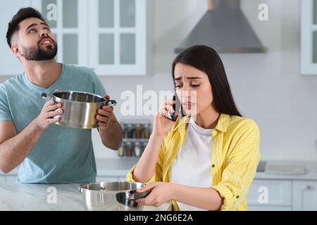 Ein junger Mann sammelt Wasser aus der Decke, während seine Freundin den Dachreparaturservice in der Küche anruft Stockfoto