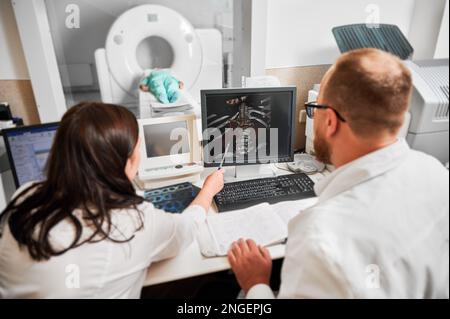 Medizinische Computertomographie oder MRT-Scanner. Rückansicht von zwei Ärzten, die am Computer sitzen und MRT-Ergebnisse untersuchen. Spezialisten Frau zeigt mit Stift, Patient lügt. Der Begriff der Medizin. Stockfoto