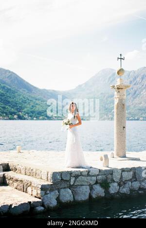 Dobrota, Montenegro - 06.06.21: Die Braut mit einem Blumenstrauß steht auf einem Pier neben einer Säule mit einem Kreuz Stockfoto