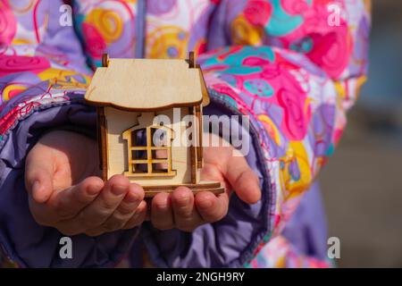 Die Kinderhand hält ein kleines Holzhaus auf einem verschwommenen Hintergrund Stockfoto