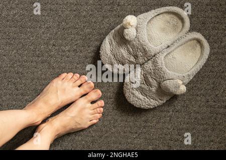 Weibliche Füße und graue, weiche Hausschuhe auf grauem Teppich, Innenschuhe aus nächster Nähe Stockfoto