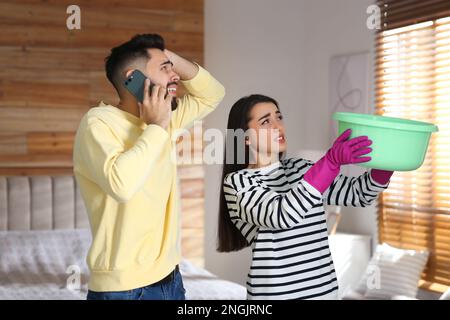 Eine junge Frau sammelt Wasser von der Decke, während ihr Freund zu Hause den Dachreparaturservice anruft Stockfoto
