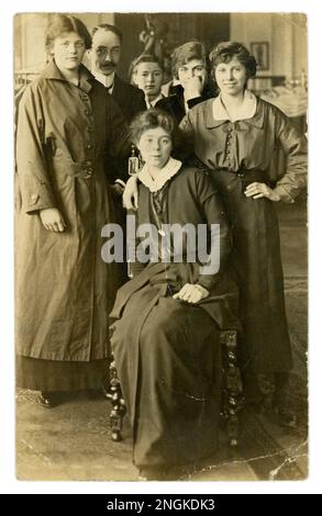 Originale Postkarte aus dem WW1. Jahrhundert mit attraktiven, jungen Frauen, möglicherweise Freunden oder Schwestern - eine trägt Arbeitskleidung mit Mantel. Ein Junge und ein Mann schauen sich um, vielleicht ein Assistent, Familienfeier, Hochzeitsgruppe, in Ausstattern vielleicht als Anzüge an Kleiderbügeln oder wird das Mädchen rechts für einen Arbeitsmantel ausgestattet? Veröffentlicht von Tooting, South London, Großbritannien, datiert/veröffentlicht am 30. April 1915 Stockfoto