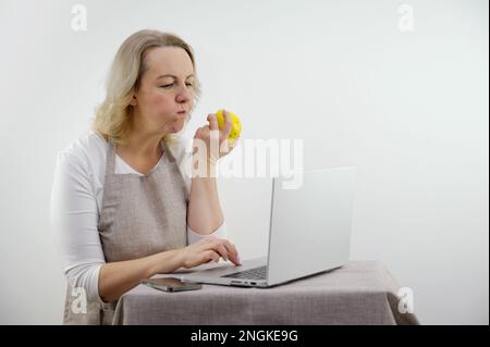 Schürze Telefon Laptop Foto einer schönen Frau, die ihr Smartphone benutzt, während sie Apfel isst, während sie zu Hause auf der Treppe sitzt. Hochwertiges Foto Stockfoto