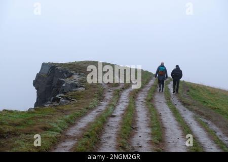 Edinburgh, Schottland, Großbritannien. 18. Februar 2023 Die Menschen gehen unter dem bleigrauen Himmel spazieren, während niedriger Nebel und hartnäckiger Nieselregen die Salisbury Crags im Holyrood Park umhüllen. Kredit: Craig Brown/Alamy Live News Stockfoto