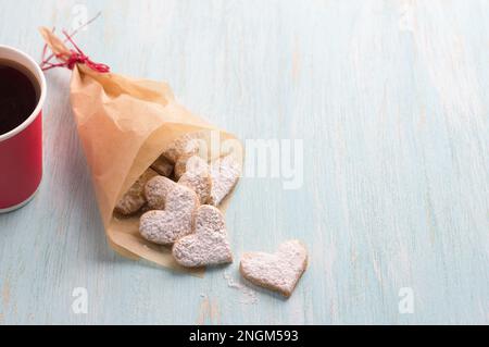 Köstliche hausgemachte herzförmige Kekse mit Puderzucker in einer handgefertigten Tüte zum valentinstag als Geschenk mit Pappbecher Kaffee auf hellblauem Ba Stockfoto