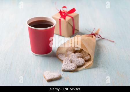 Köstliche hausgemachte herzförmige Kekse mit Puderzucker in einer handgefertigten Tüte zum valentinstag als Geschenk mit Pappbecher Kaffee auf hellblauem Ba Stockfoto