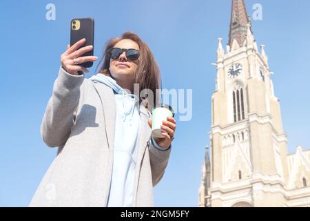 Eine Frau mit Sonnenbrille in einem Mantel, die auf dem Platz der Altstadt steht und eine Kaffeetasse und ein Handy in der Hand hält. Stockfoto