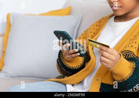 Frau kauft online mit Smartphone und bezahlt mit goldener Kreditkarte. afroamerikanisches Mädchen, das im Internet kauft, Kreditkartendetails eingeben. E-Commerce-Lieferservice für Online-Shopping Stockfoto