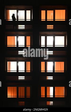 Beleuchtete Apartmentfenster in der Nacht im städtischen Gebäude mit der Silhouette einer Person am Telefon oder Raucher auf dem Balkon in vertikaler Form Stockfoto