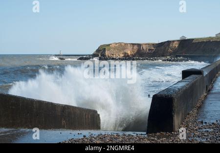 Sehen Sie entlang der Promenade von Seaham, einschließlich Klippen und entferntem Leuchtturm, mit Wellen, die in die Steinmauern stürzen und Spritzer auswerfen Stockfoto