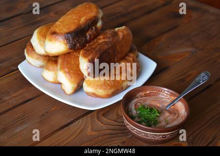 Gebratene Würstchen in Teig auf dem Teller mit herzhafter Tomatensoße und frisch gehacktem Dill in der kleinen Tonschüssel auf dem Holztisch. Stockfoto