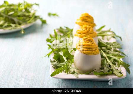 Gekochte Eier gefüllt mit gebratenen Zwiebeln und Kurkuma mit frischem Rucola auf hellblauem Hintergrund. Köstlicher hausgemachter Snack Stockfoto