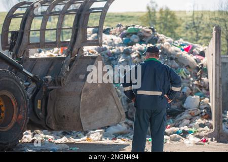 Ein Arbeiter im Recycling-Center, in dunkelblauer Arbeitskleidung, der sich einen unsortierten Müllhaufen ansieht, ist gerade in der Materialrückgewinnungsanlage angekommen, Rückansicht. Stockfoto