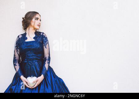 Wunderschönes Mädchen in glamourösem, ultramarinem Kleid, das an der Wand steht, mit einer herzförmigen Handtasche Stockfoto