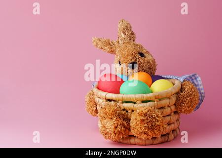 Korb mit Häschen aus Stroh mit bunten ostereiern auf pinkfarbenem Hintergrund. Frohe Ostern. Speicherplatz kopieren. Schließen Stockfoto