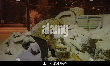 Der vom Militär bekleidete junge Mann, der den harten Winter überlebt, hackt Holz mit Axt, um sich bei schneebedeckten Bedingungen warm zu halten Stockfoto