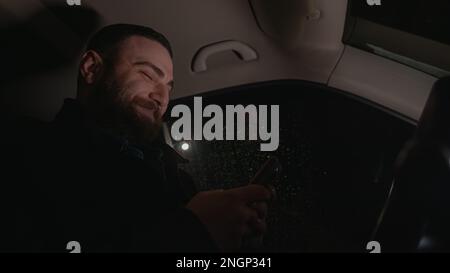 Selbstbewusster und glücklicher junger Mann mit einem bärtigen Gesicht, der in einem Auto auf seinem Telefon schickt, sich amüsiert und unterwegs mit dem Convenien kommuniziert Stockfoto