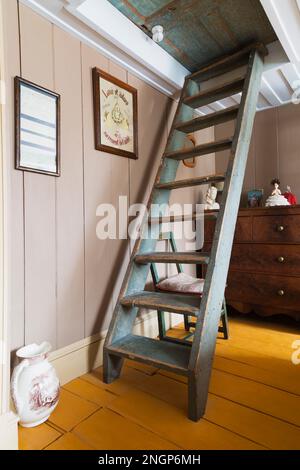 Hölzerne Müllertreppe führt zu Dachboden und Kommode im oberen Gästeschlafzimmer mit gelb bemalten breiten Brettern im alten 1835-Haus. Stockfoto