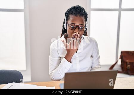 Afrikanische Frau mit geflochtenem Haar, die ein Headset für Callcenter-Agenten trägt, das den Mund mit der Hand verdeckt, schockiert und vor Fehlern fürchtet. Überraschter Ausdruck Stockfoto