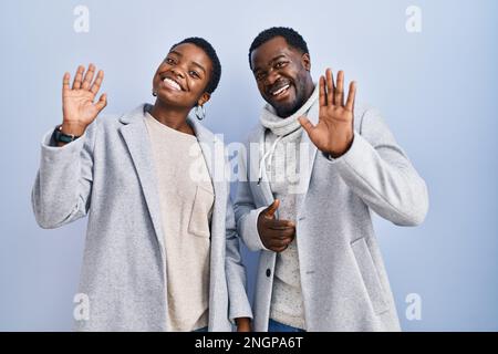 Ein junges afroamerikanisches Paar, das auf blauem Hintergrund steht und darauf verzichtet, Hallo zu sagen, fröhlich und lächelnd, freundliche Willkommensgeste Stockfoto