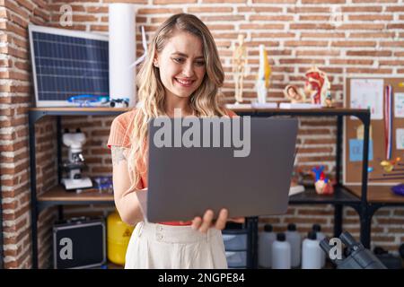 Junge blonde Lehrerin, die selbstbewusst mit dem Laptop im Klassenzimmer lächelt Stockfoto