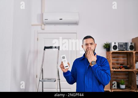 Hispanischer Mechaniker, der mit Klimaanlage arbeitet, verdeckte den Mund mit der Hand, schockiert und Angst vor Fehlern. Überraschter Ausdruck Stockfoto