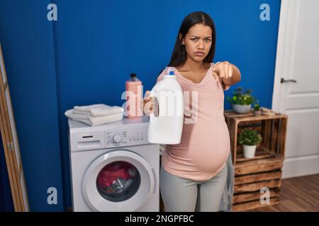 Junge schwangere Frau, die Wäsche wäscht und die Waschmittelflasche hält und mit dem Finger auf die Kamera und auf dich zeigt, selbstbewusste Geste, die ernst aussieht Stockfoto