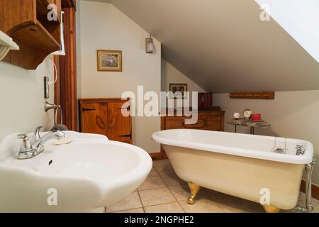 Weißes Waschbecken mit Sockel, Badewanne mit Klauenfuß und antiken Kiefernholzschränken im Bad mit Keramikfliesen auf der oberen Etage im 1990er modernen V Stockfoto