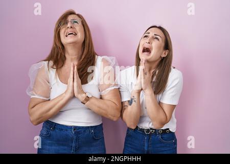 Hispanische Mutter und Tochter tragen ein legeres weißes T-Shirt auf pinkfarbenem Hintergrund und betteln mit den Händen zusammen mit dem Ausdruck der Hoffnung im Gesicht Stockfoto