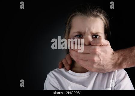 Ein Mann, der den Mund eines verängstigten kleinen Mädchens auf schwarzem Hintergrund verdeckt, Platz für Text. Häusliche Gewalt Stockfoto