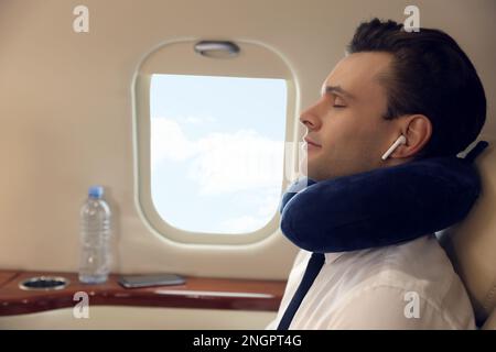 Junger Mann mit Reisekissen ruht sich aus, während er während des Fluges Musik im Flugzeug hört Stockfoto