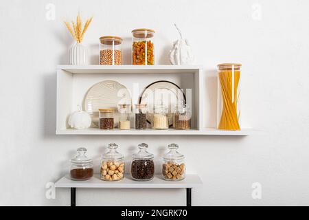 Ein Set aus Glas- und Keramikschalen, glasgefüllte Gläser mit Großprodukten auf weißen Küchenregalen an strukturierten Wänden. Kompakter Stauraum. Elegantes Design Stockfoto