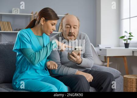 Junge Pflegekraft, die zusammen mit einem älteren Mann auf dem Sofa sitzt und ihm hilft, sein Smartphone zu benutzen Stockfoto
